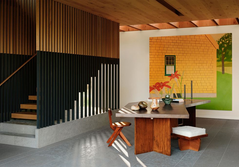 entryway with gray tile floor, a table with wood base and angular top, vases, objects, candles on top, a chair and stool, large colorful artwork, concrete stairway with wood steps and black and wood floor to ceiling balustrade