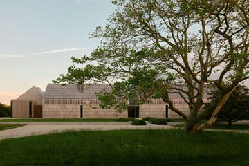 a long modern house with a side building made of cedar shingles and pitched roof, grass with a large tree, a gravel driveway and walkway with shrubs to house