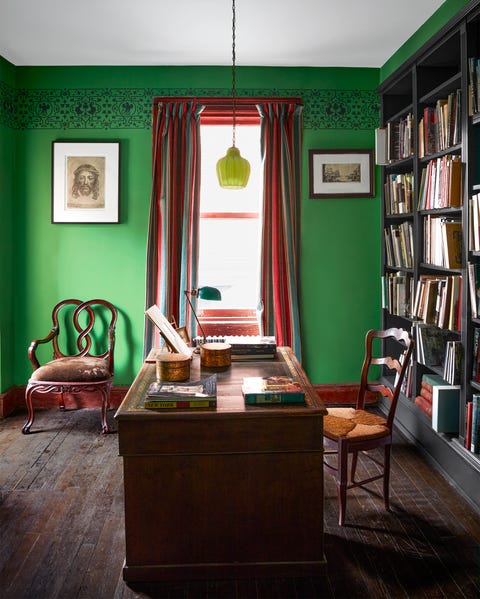 an antique wooden desk and chair sit in front of floor to ceiling book shelves, a green glass pendant is above desk, the walls are green with a painted ornamental band near the ceiling, and there is a curtained window