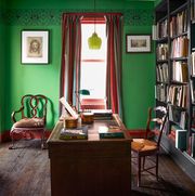 an antique wooden desk and chair sit in front of floor to ceiling book shelves, a green glass pendant is above desk, the walls are green with a painted ornamental band near the ceiling, and there is a curtained window