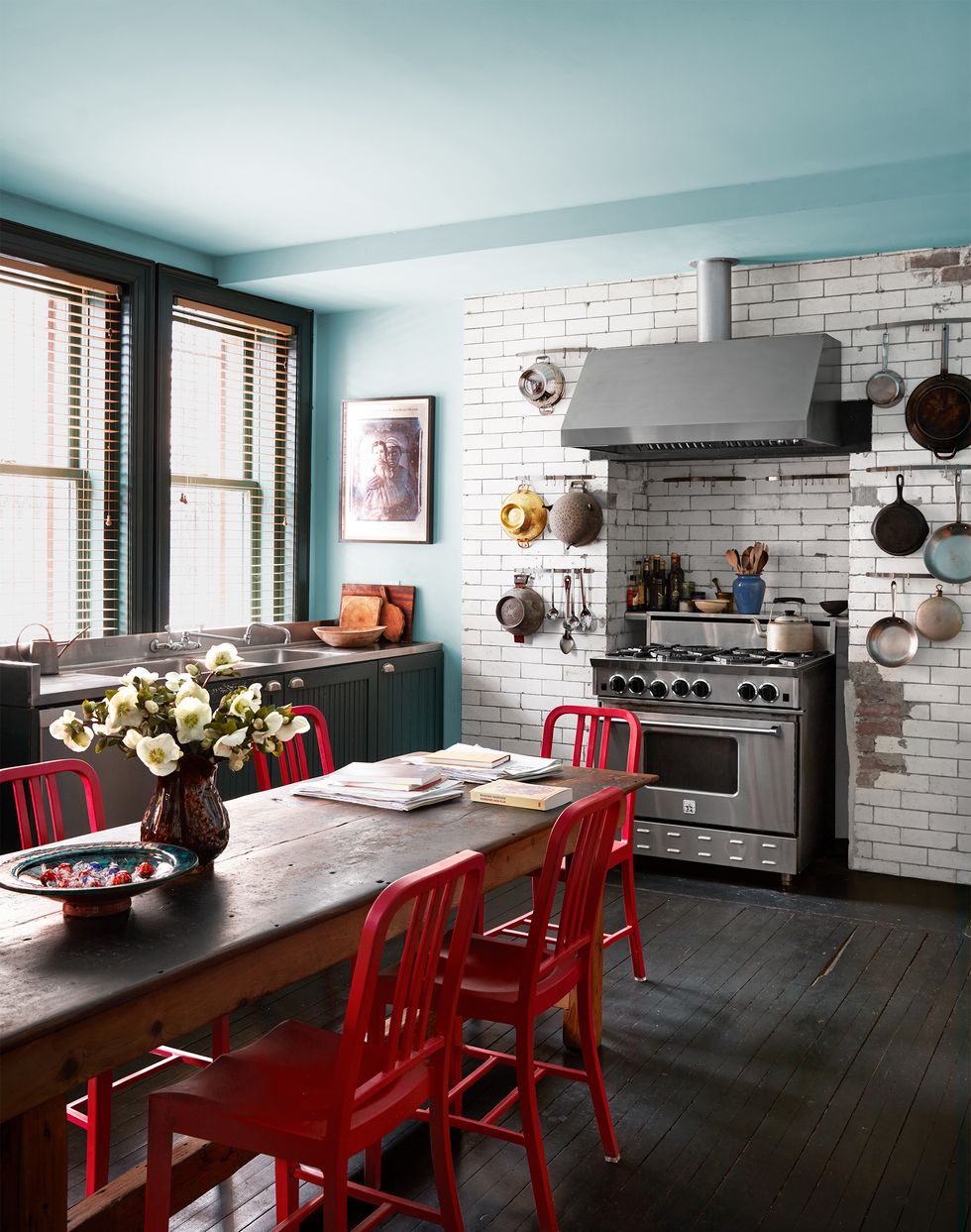 a kitchen has a long antique wood table with red chairs, a dark wood floor, light blue walls and ceiling, subway tiles surrounding a stove and pots and colanders hanging on both sides of it