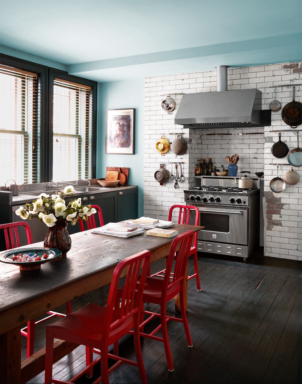a kitchen has a long antique wood table with red chairs, a dark wood floor, light blue walls and ceiling, subway tiles surrounding a stove and pots and colanders hanging on both sides of it