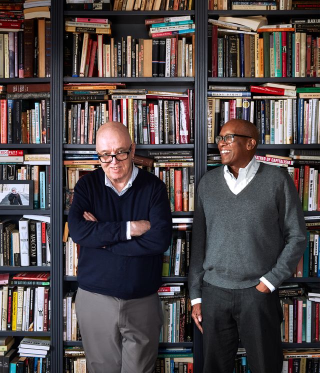 the two homeowners, both wearing eyeglasses, stand in front of floor to ceiling bookshelves