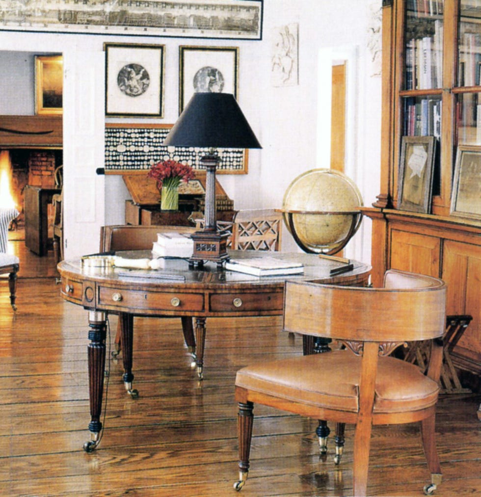 cozy room with round regency table with small drawers around it and on wheels with antique round back chairs and a lamp with a black shade on it and a wood hutch on the right and view of the living room with fireplaced in the far background