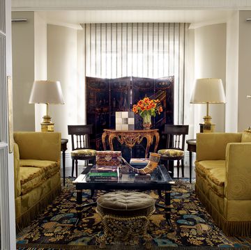 elegant living area seen through portieres with facing velvet yellow sofas and matching white lamps with gold bases and a dark cocktail table at center over a patterned dark rug and small tufted greenish brown stool and a mahogany screen at back in front of a window