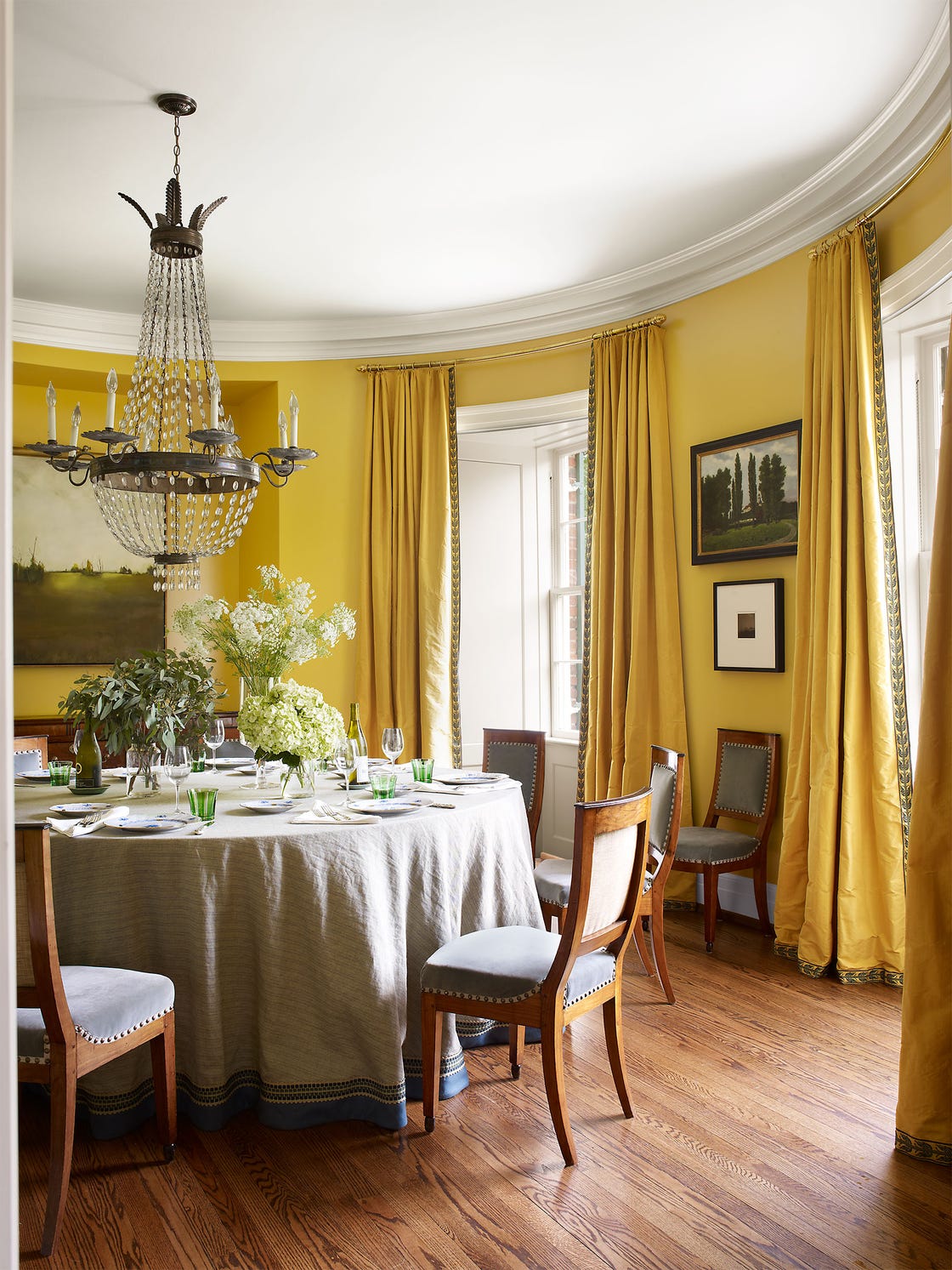 round yellow dining room with wood floors a chandelier and chairs and table with floor length tablecloth and yellow curtains on tall windows