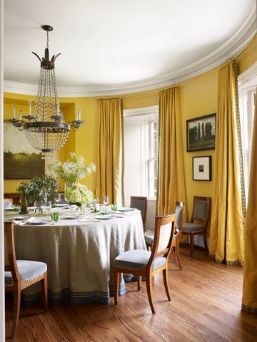 round yellow dining room with wood floors a chandelier and chairs and table with floor length tablecloth and yellow curtains on tall windows