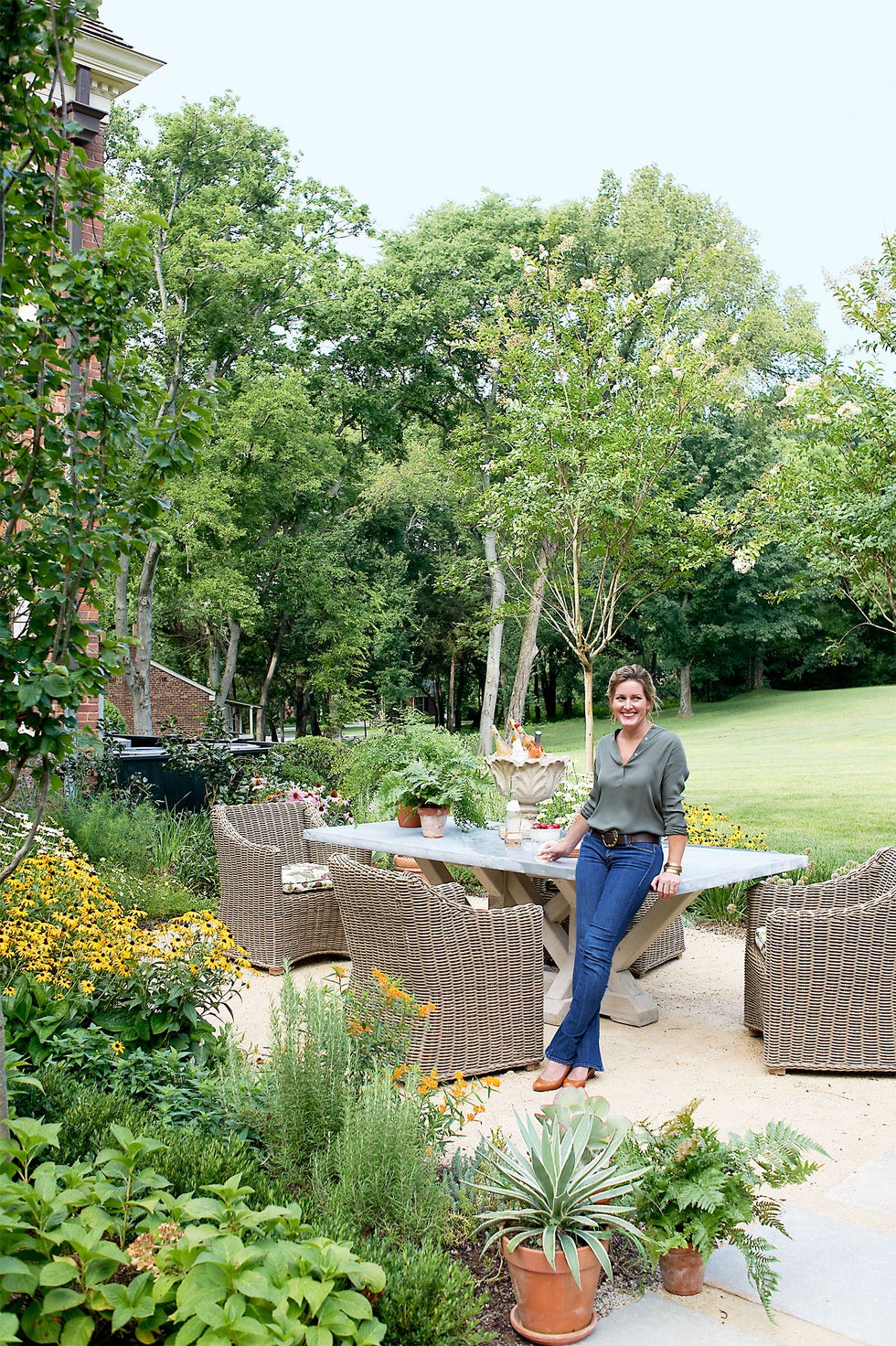 woman in jeans leaning on a pedestal marble table outside surrounded by wicker chairs and lots of plans and trees