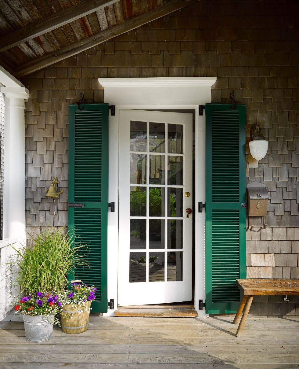 white pained front door with gorgeous full height green shutters and a bench on the right and some potted plants on the left and the decking is wooden planks