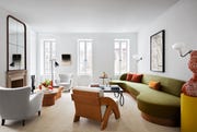 a living room has three windows, white walls, a fireplace with carved mantel and large mirror above, two armchairs, a lounge chair, two cocktail tables, a long green curved sofa, wall sconces, and artworks