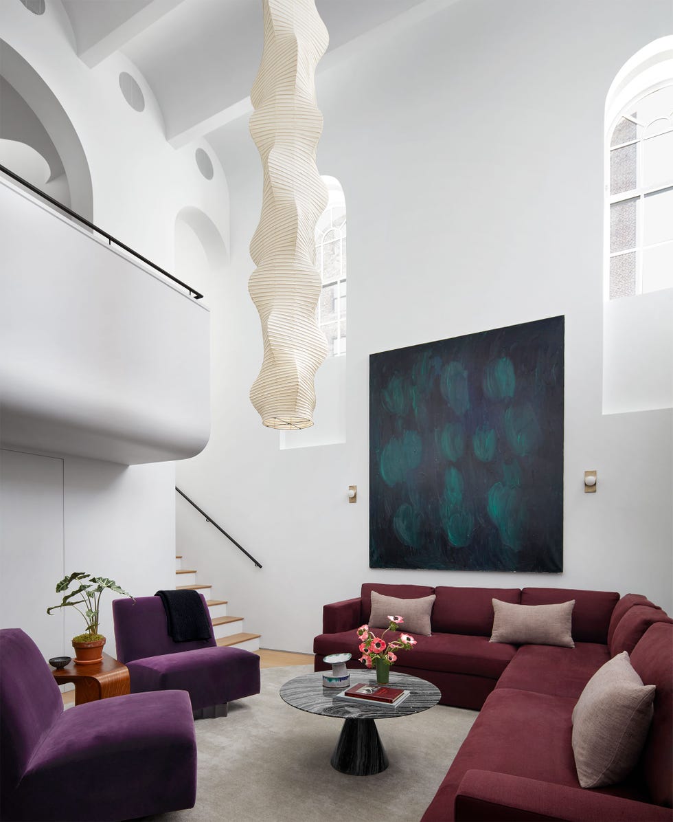 Living area with high ceilings, dark burgundy wraparound sofa with dark purple velvet armless chairs and a low coffee table with triangular base and large pendant hanging from the ceiling