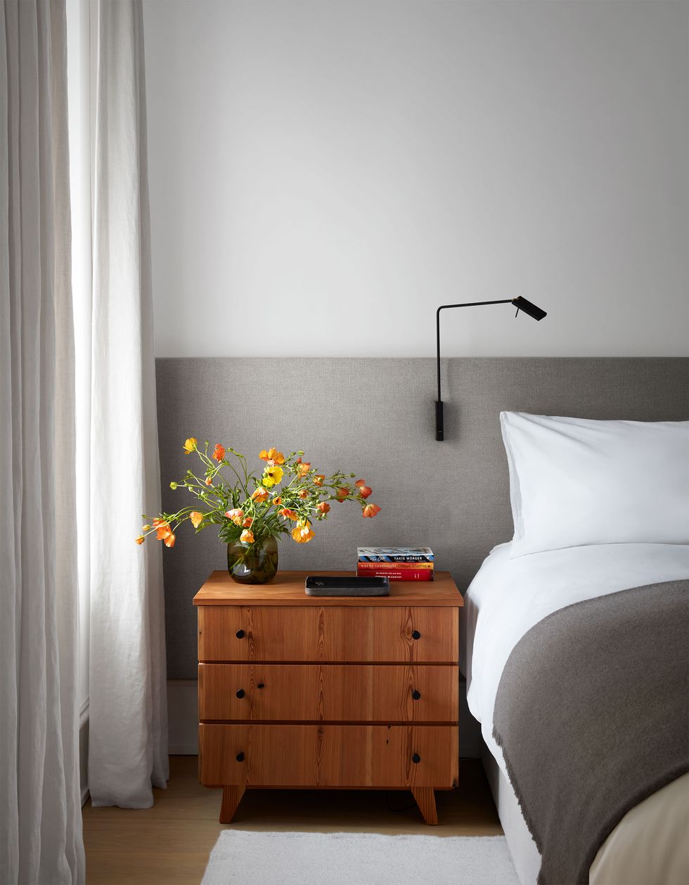 a bed has an oatmeal fabric headboard that extends along the wall with a reading sconce attached, a side table with three drawers and books and a vase with flowers atop it, curtains hang on the window