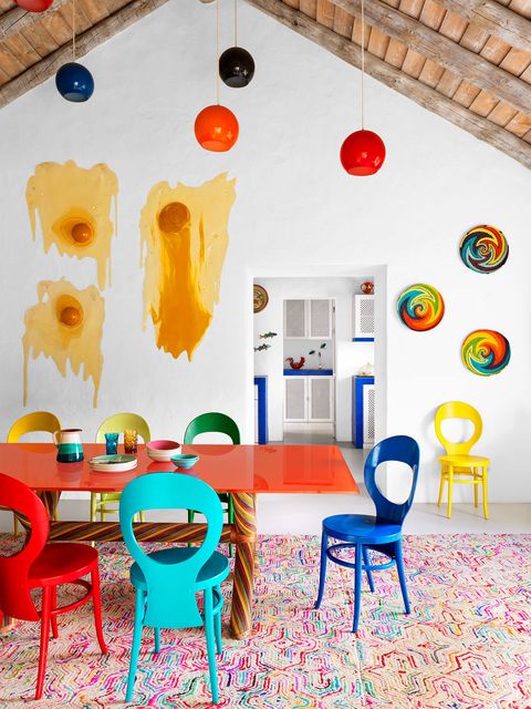dining table topped with orange glass with woven rattan legs and bright painted chairs in different colors, multicolored patterned rug, five pendant lights, swirled pattern bright baskets and resin art hanging on wall