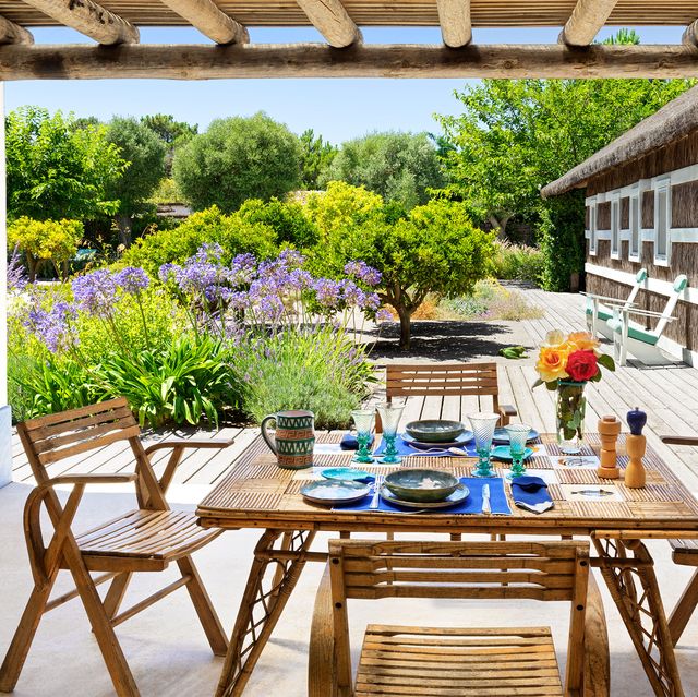 a covered terrace outside a wing of the house has a square bamboo dining table with four chairs, a deck with lounge chairs and small trees, tall grasses, and bushes with lavender colored blossoms