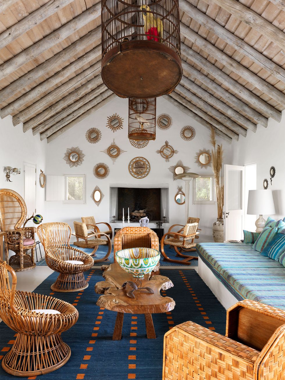 A living room with a birdcage hanging from the diagonal beam ceiling, a fireplace with some straw mirrors on the other side of the wall, wicker chairs, a rustic wooden cocktail table, a striped light blue fabric sofa and a small red square. navy rug