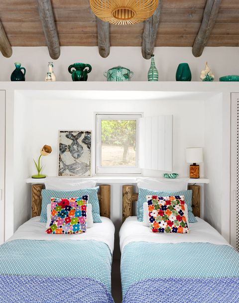 a guest room has a small window and two twin beds with rustic wood frames, aqua printed blankets and blue printed throws and several pillows, a shelf at head of beds has two lamps and an artwork with fish images