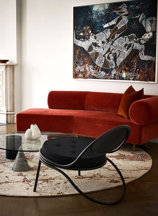 living area with a rolled arm chaise covered in red velvet, a low black metal armless lounge chair with fabric cushion, round glass cocktail table, round light patterned rug, collage like abstract artwork above sofa