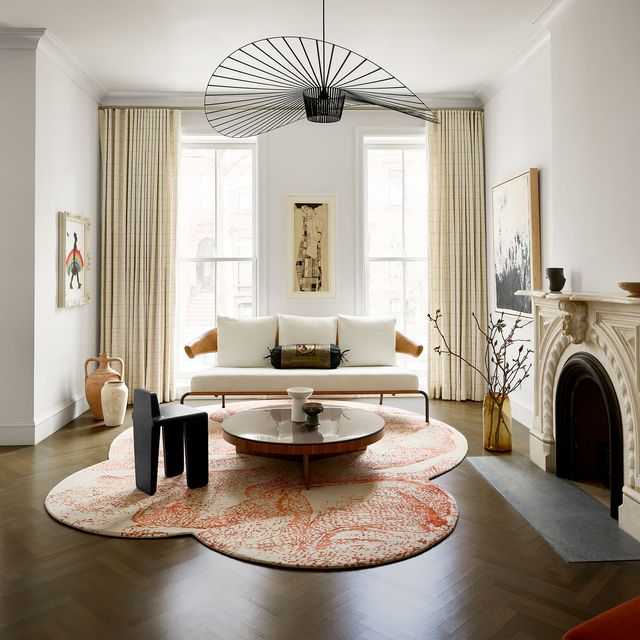 living room with floor to ceiling windows with drapes, a wood and metal framed sofa with white cushions, round cocktail table, dark wood stool, flower petal shaped rug, carved stone fireplace, a hoop shaped pendant light