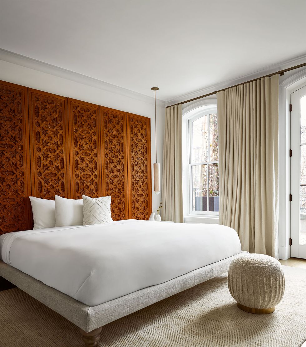 The master bedroom has a wood and linen fabric bed frame and white linens, a Moorish carved wood door forms a headboard along the back wall of the bed, curtains, a beige pouf Man, the rug is beige