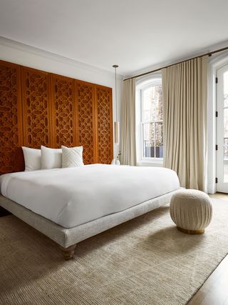 primary bedroom has a wood and linen fabric bed frame with white linens, moorish carved wood doors line the wall behind bed forming a headboard, the drapes, a beige pouf, and the rug are beige colored
