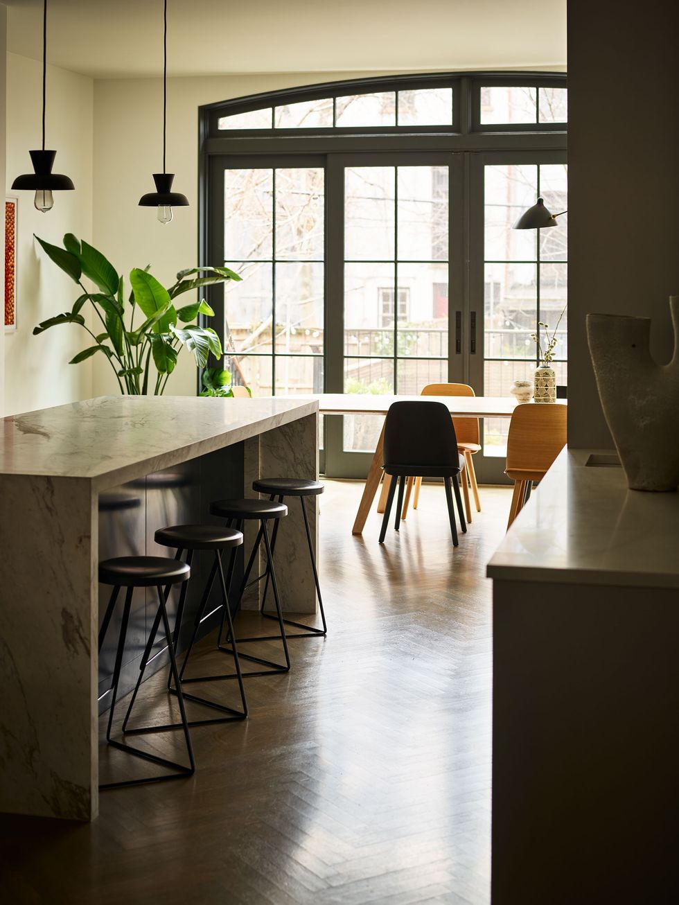 sliding glass doors let line into a kitchen that has a dining table and wooden chairs, a marble counter with four backless stools and two pendants above, a wood floor, and a large potted palm in the corner