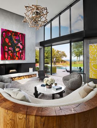 a living room with a glass wall looks out onto a deck and pool and has a sofa with a thick wood frame and light nubby fabric, upholstered armchairs, a round white glass cocktail table, a chaise, a fireplace, and large artworks