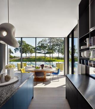 a kitchen has a glass wall overlooking a pool, steel blue floor cabinets with a terrazzo counter across from dark wood cabinets with a steel counter with shelves above, a round table with different colored woven fabric chairs