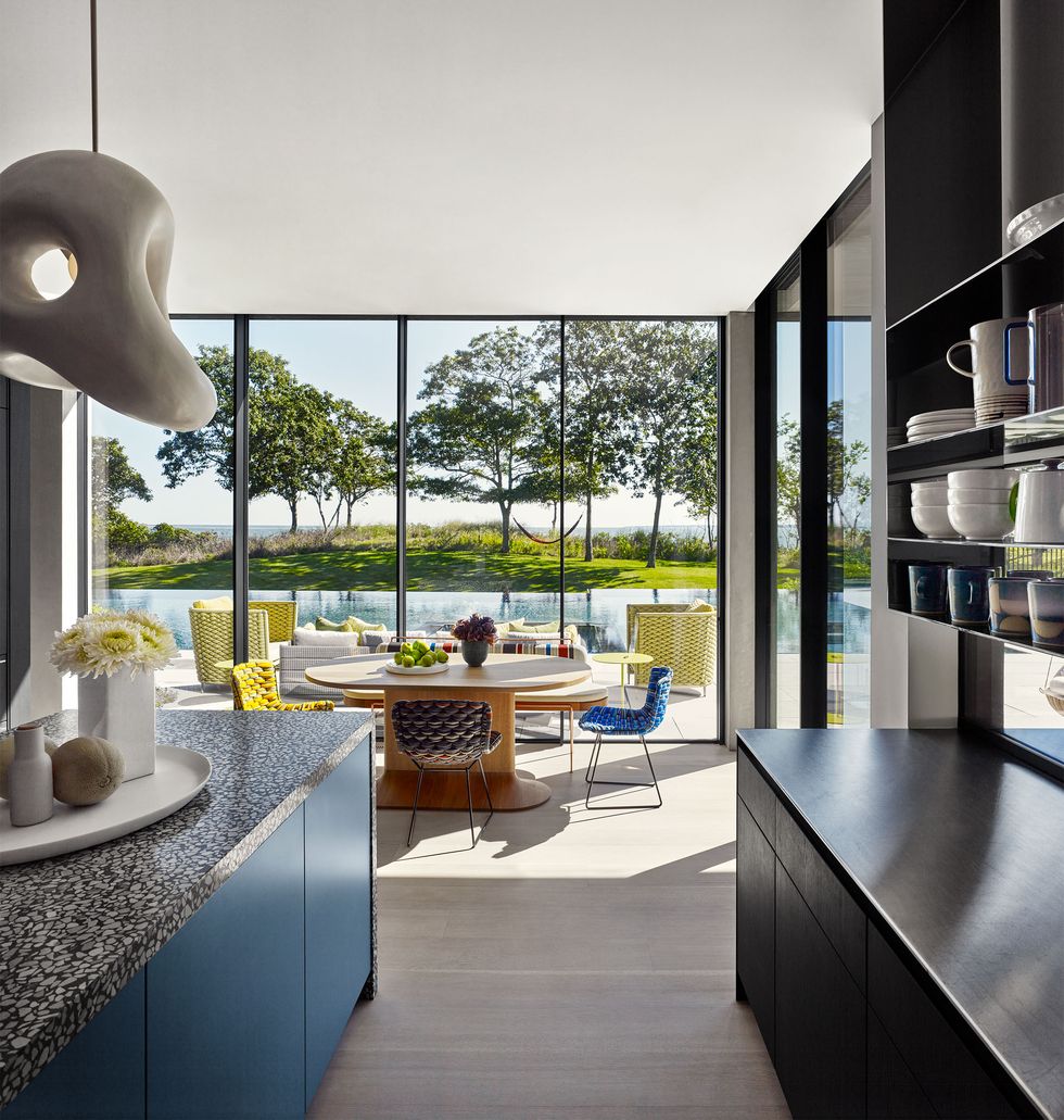 a kitchen has a glass wall overlooking a pool, steel blue floor cabinets with a terrazzo counter across from dark wood cabinets with a steel counter with shelves above, a round table with different colored woven fabric chairs