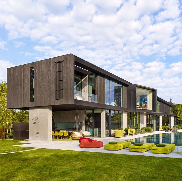 the exterior of the two story house with charred wood cladding on the upper floor and glass walls and concrete on the lower looks out onto a large swimming pool and deck with bright green, pink, and orange lounge chairs