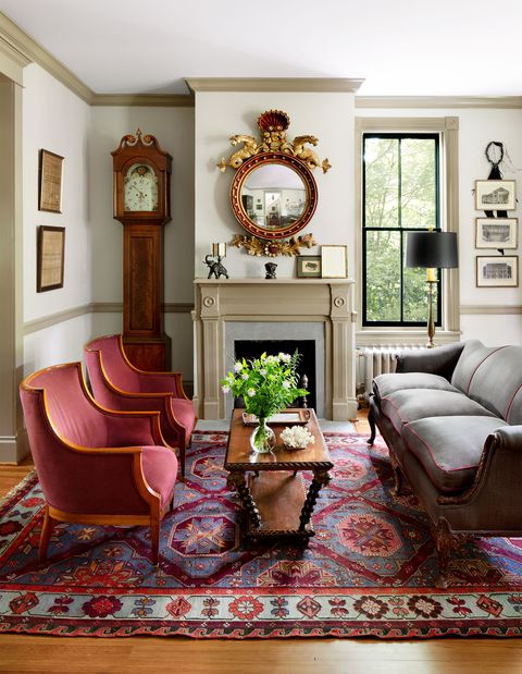 in a living is a fireplace with carved wood mantel and an ornate round mirror, a grandfather clock, two armchairs with burgundy upholstery, a cocktail table with turned legs, a gray sofa, and a patterned rug