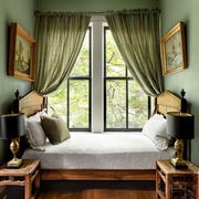 tucked into a curtained window nook is a fruitwood daybed with turned urn finials and decorative pillows, two antique chinese side tables each with a bronze lamp, and framed landscape paintings on facing walls
