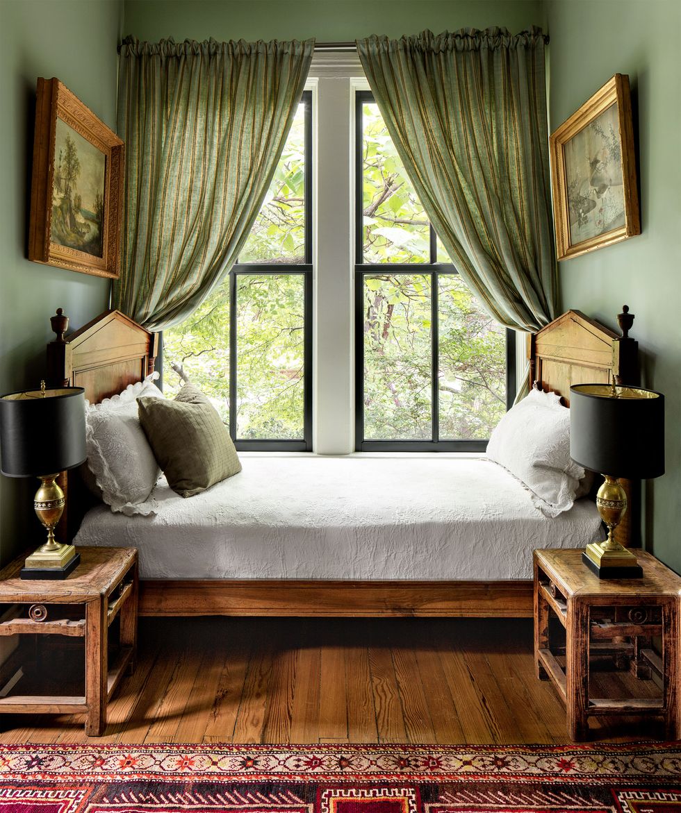 tucked into a curtained window nook is a fruitwood daybed with turned urn finials and decorative pillows, two antique chinese side tables each with a bronze lamp, and framed landscape paintings on facing walls