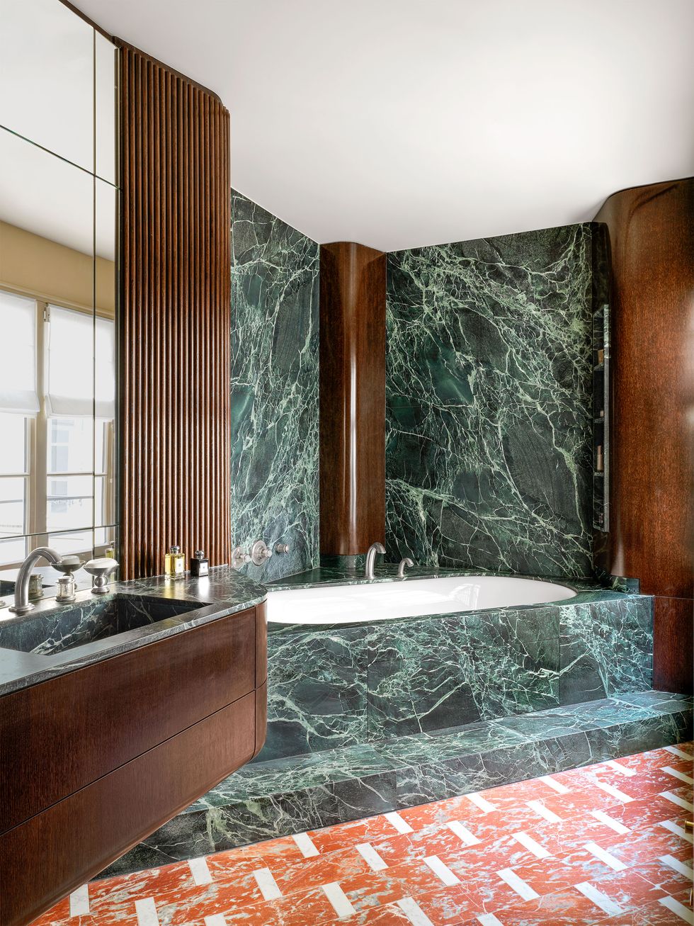 a bathroom has dark wood vanity with a sink at left against a mirrored wall, a bathtub encased in dark green marble with dark wood accented walls, the floor is red and white marble
