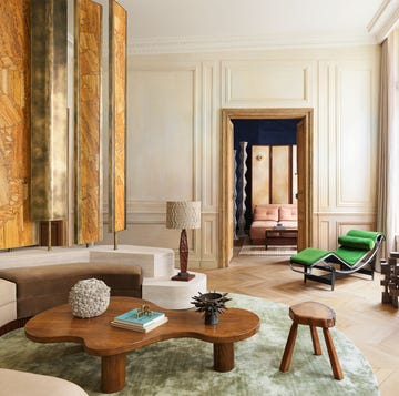 living room with beige armless sofa and four decorative rotating yellow travertine panels on a platform behind it, a curvy wooden cocktail table, a vintage lamp, stool, a bright green chaise, and a muted green rug