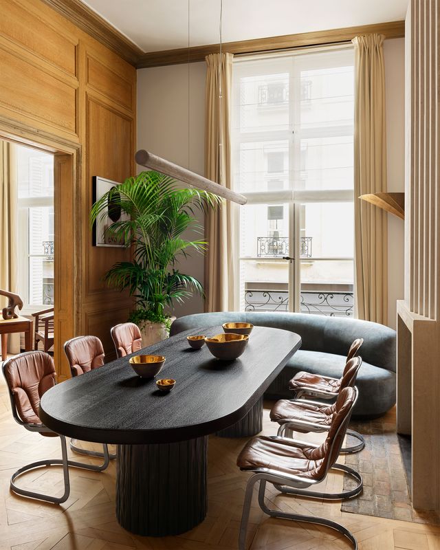 in a dining area a long light fixture hangs over a long dark wood oval table supported by pedestals with three leather and metal chairs on each side and a curved dark green velvet banquette at the end in front of a window