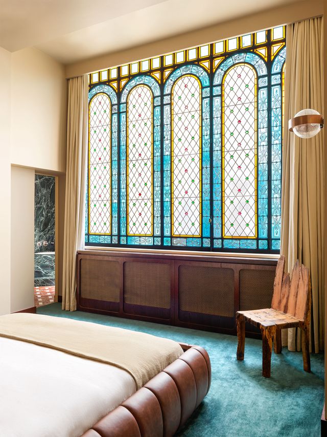 a bedroom dominated by a colorful stained glass window with a pattern of large arches, lattice, and small squares, has a quilted leather bed frame has off white and beige bedding, a green carpet, and rustic chair