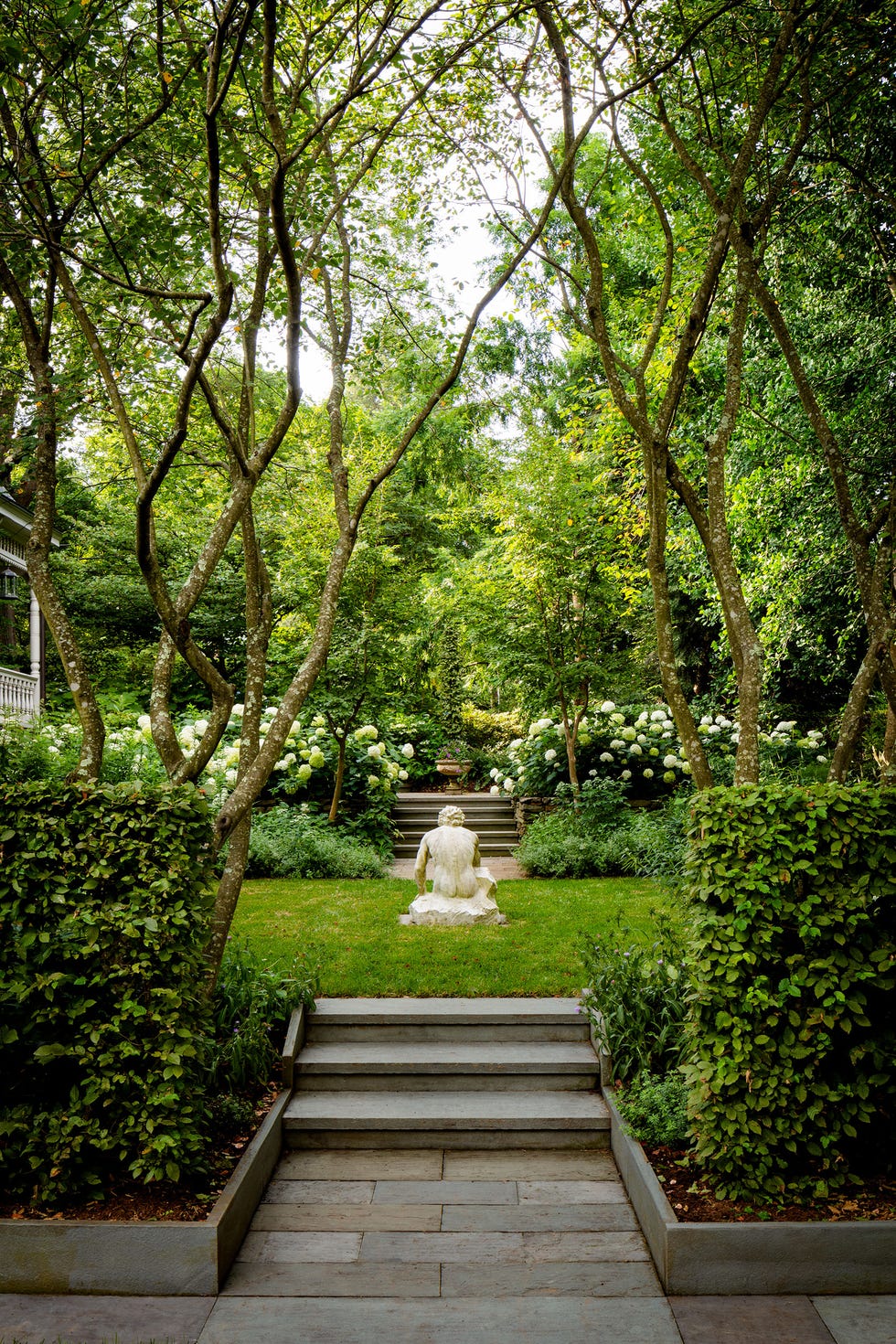 stone steps with bushes on either side lead up to a grassy clearing with a marble sculpture of a person seated on a stone, with more steps and blooming hydrangeas on the far side