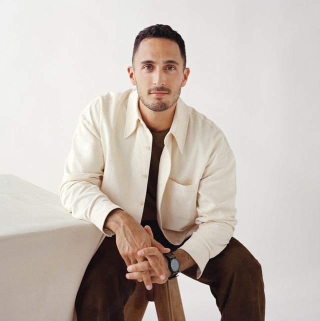 designer in a white shirt and brown pants leaning on a block table looking into the camera