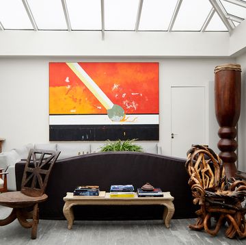 a white living room with a gray floor has a skylight ceiling, a painting of a matchstick, a brown sofa, a white sofa, a wooden african chair, a chair made of mangrove roots, a tall drum from the philippines