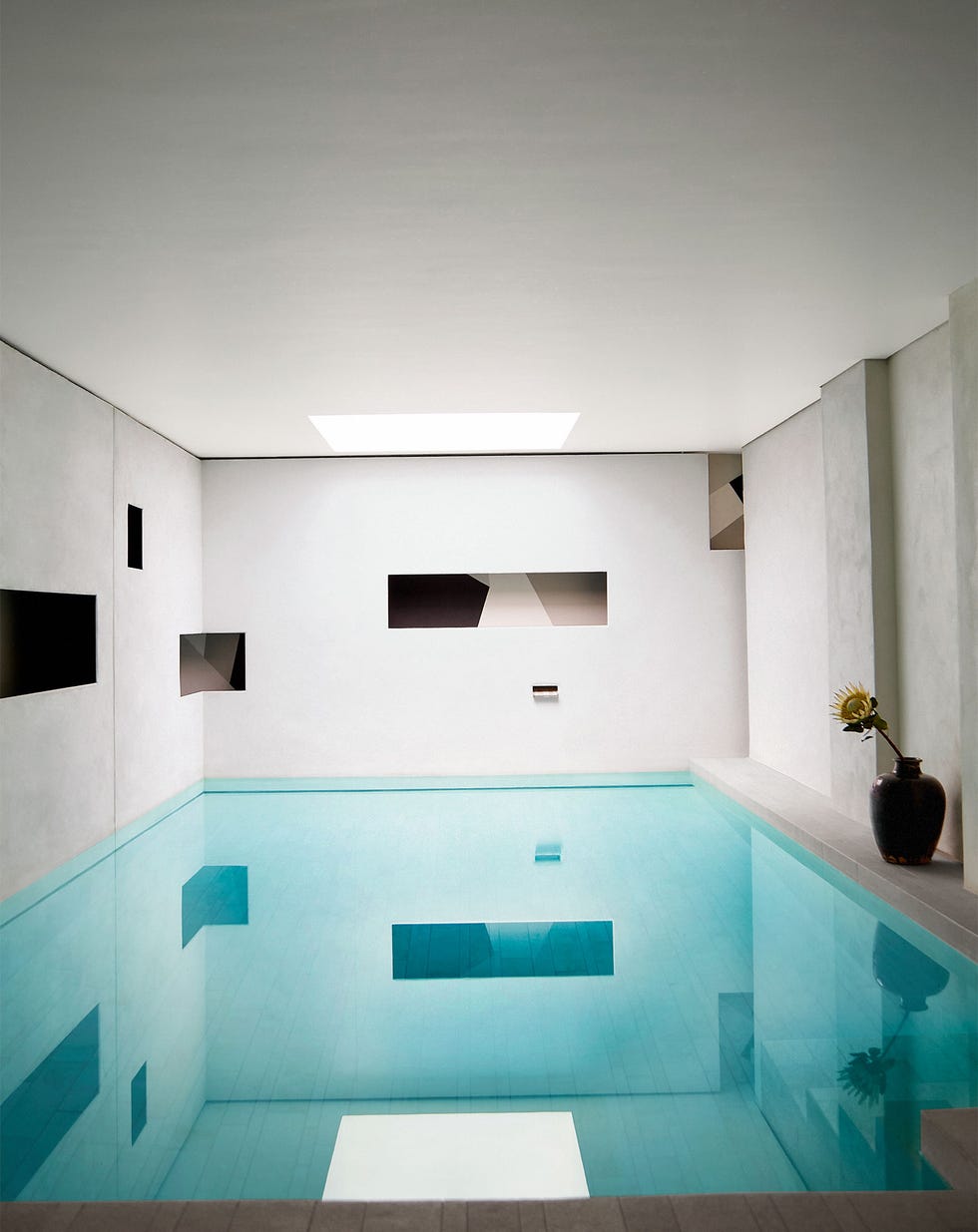 in a basement is a full room pool made with beige tiles on sides and bottom, walls have zero edges opening to a black and white fresco, a vase with a sunflower is against the wall, and a skylight is above