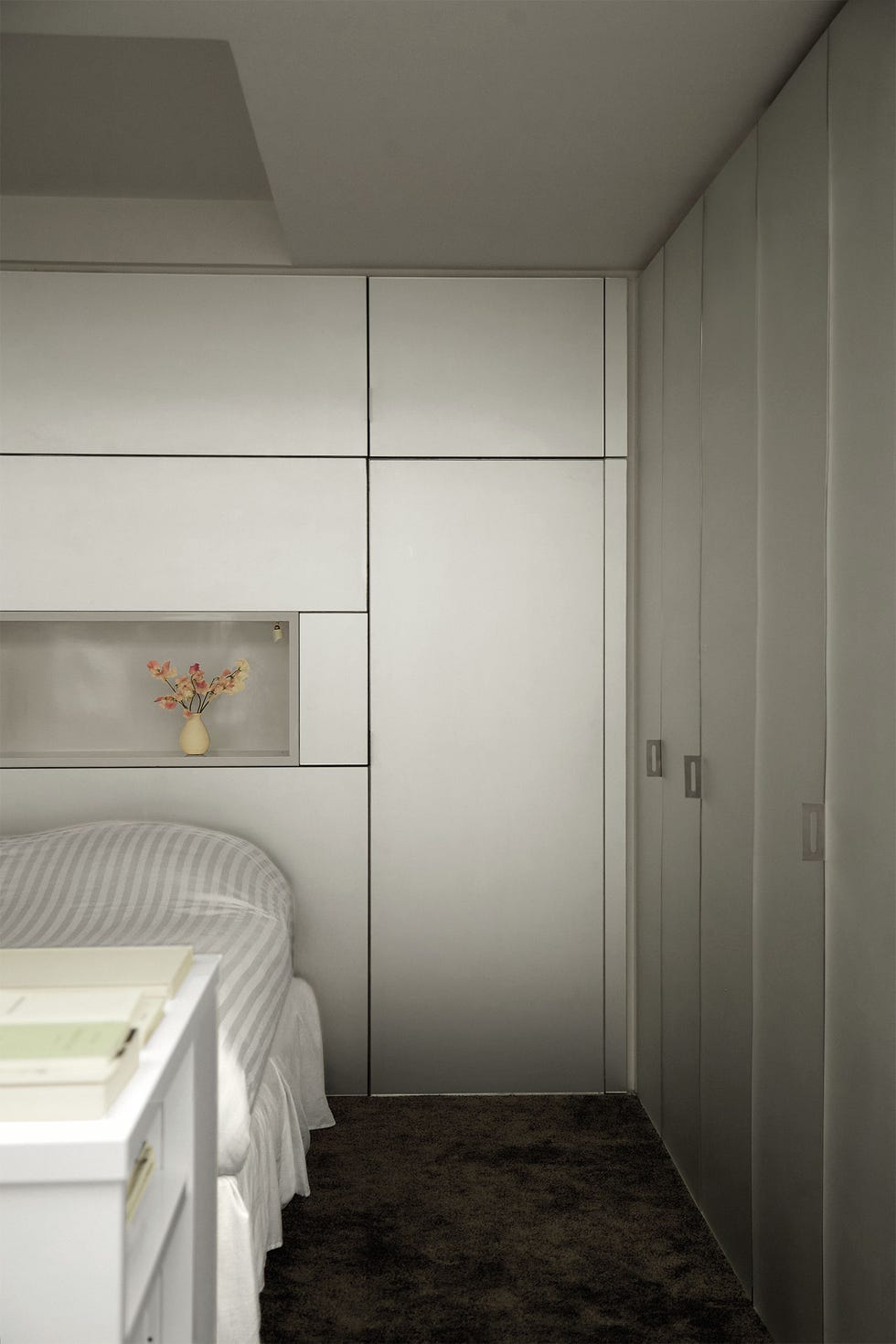 Elliott Barnes - ELLE DECOR a small bedroom has brushed steel on walls and on storage cabinet doors, a bed with a gray and white striped cover, white shelves at its foot, and an inset shelf above head of bed with a small vase and flowers