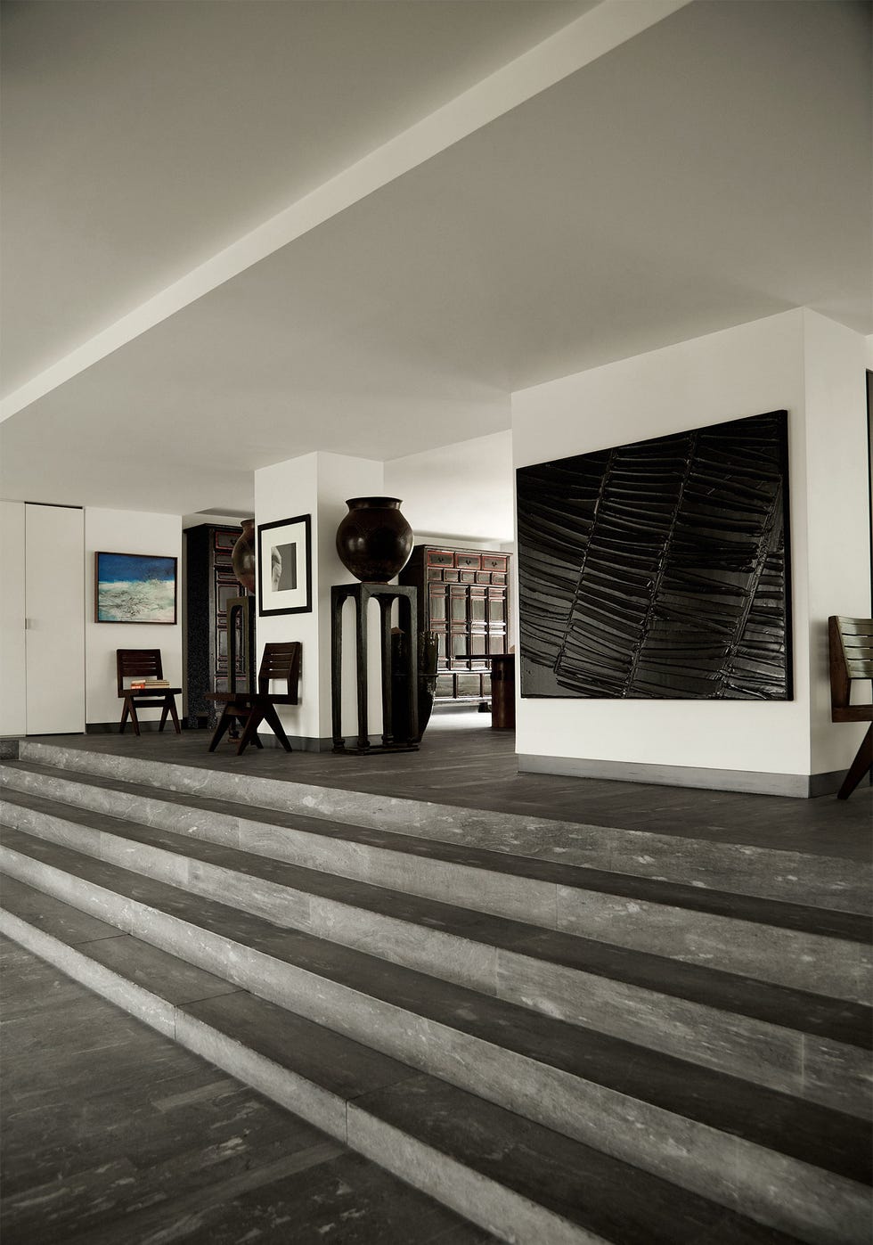 in an white walled entryway with a gray stone floor are five steps going up wide columns with paintings and photos on them, antique jars on tall plinths, vintage wooden chairs, and an antique cabinet