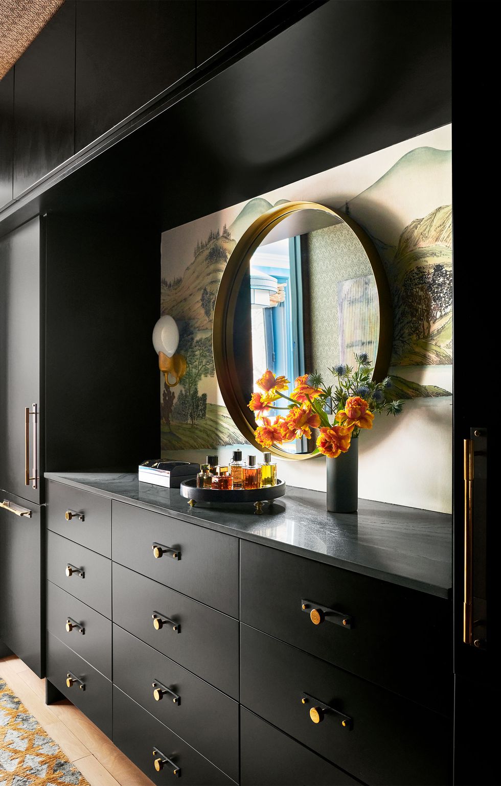 a dressing room has built in black wood cabinets and drawers which have a dark gray marble top, between upper cabinets and drawers is a round mirror hung against an asian style mural and white glass sconces