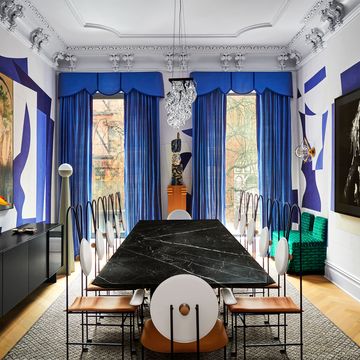 a marble dining table is surrounded by metal chairs with a leather seat and round back, blue drapes flank a sculpture, wallpaper with blue shapes, console with artwork on wall above it, crystal chandelier
