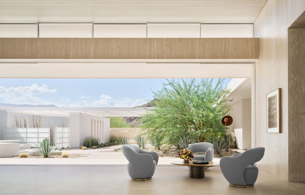 Nicole Hollis - ELLE DECOR in a desert home, three gray swivel chairs surround a round cocktail table in the room that has an entire wall open to the outdoors, where there's a courtyard with a trellis over a desert and rock garden with cacti