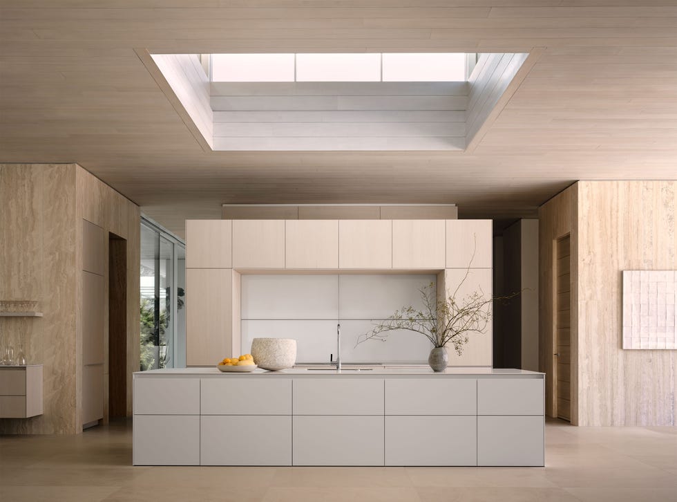a light colored monochrome kitchen has a wall of cabinets and an island with a sink and a counter with vases and a bowl of oranges, hallways on either side of the island, a large square skylight, and limestone floors