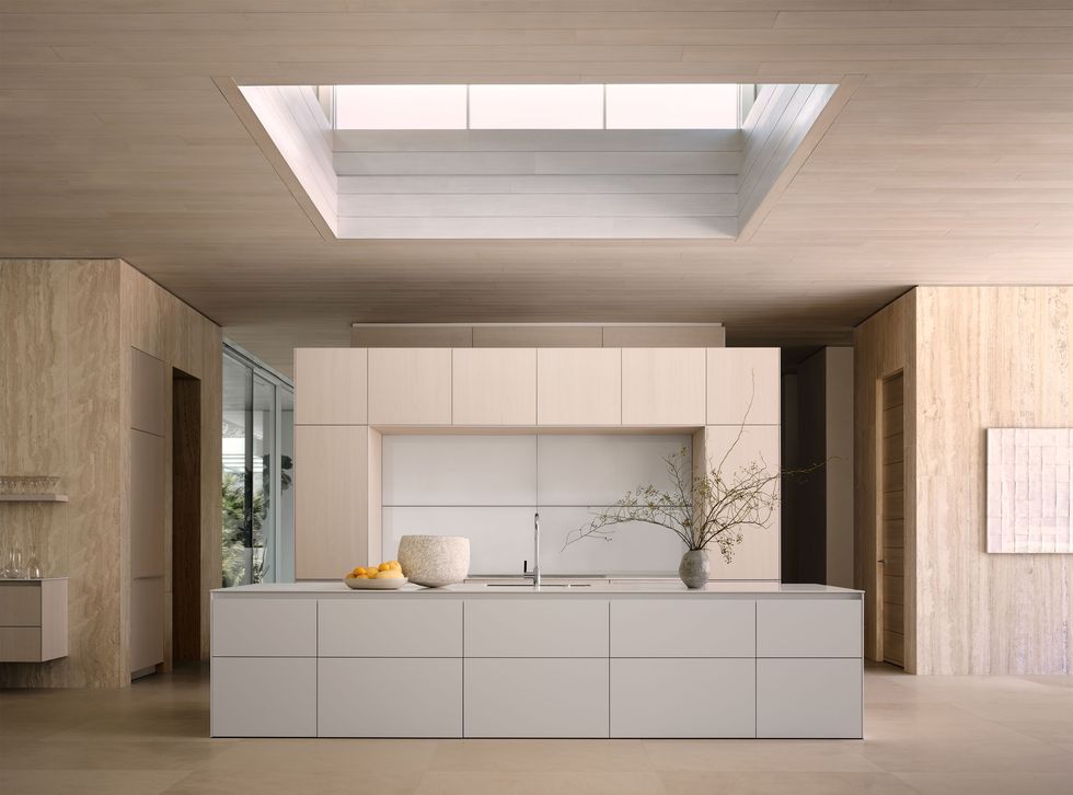 a light colored monochrome kitchen has a wall of cabinets and an island with a sink and a counter with vases and a bowl of oranges, hallways on either side of the island, a large square skylight, and limestone floors