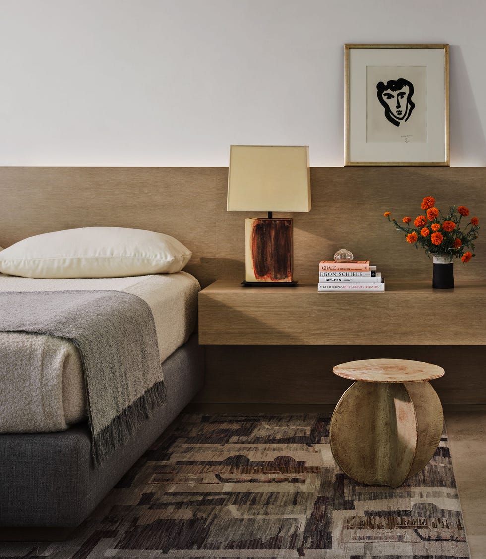 Nicole Hollis - ELLE DECOR a guest room has a bed with a connected wooden headboard and floating nightstand with a ceramic lamp, books, and a small vase on it, a ceramic stool, multitoned rug, small framed artwork by matisse