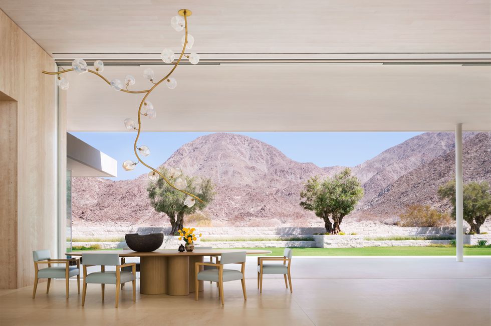 by a completely open wall looking out to a desert and mountain landscape is a large wooden pedestal table surrounded by six wooden chairs with light blue fabric seats and backs, a vinelike chandelier hangs over table