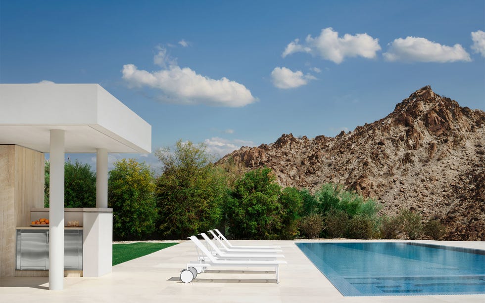 Nicole Hollis - ELLE DECOR a covered outdoor kitchen is next to a zero edge pool with a large deck and four white lounge chairs that have back wheels, beyond the pool is grass, shrubs and trees, and a rocky mountain